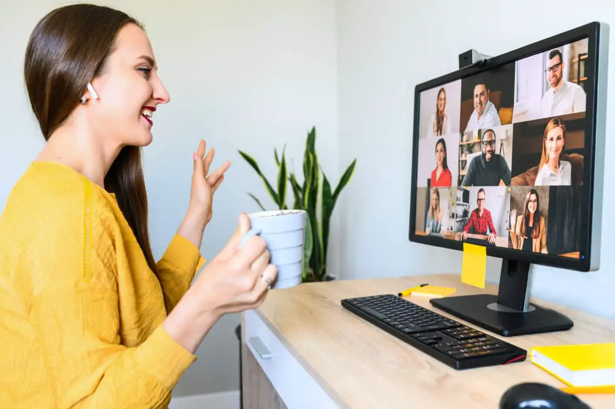 Video chat, virtual morning meeting. A young woman with cup of coffee is greeting and waving to multiracial team on the PC monitor. Side view