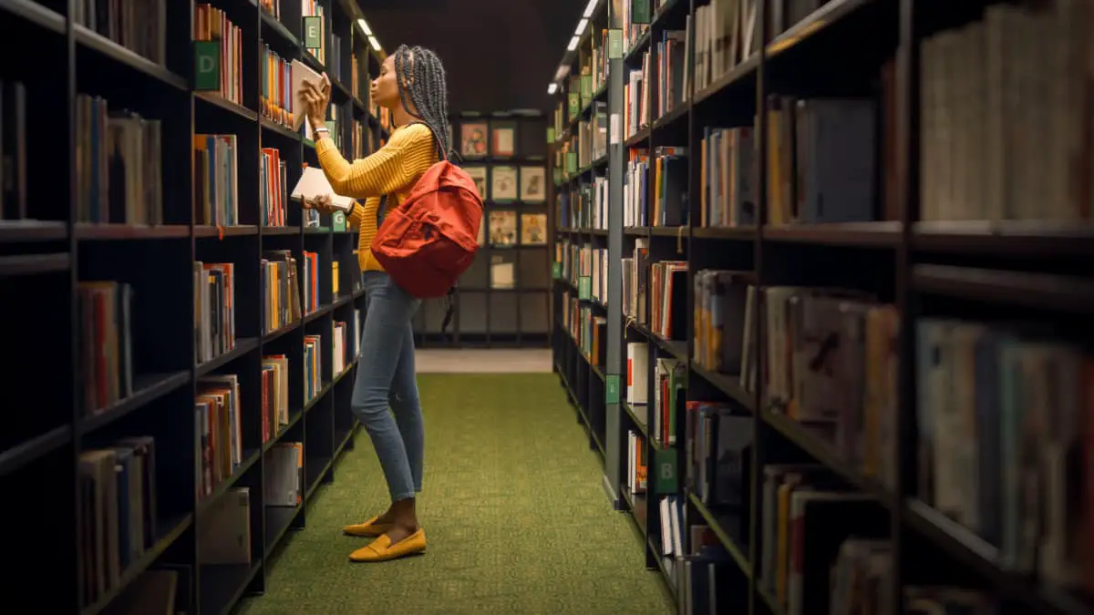 University Library: Portrait of Gifted Beautiful Black Girl Stands Between Rows of Bookshelves Searching for the Right Book for Class Assignment