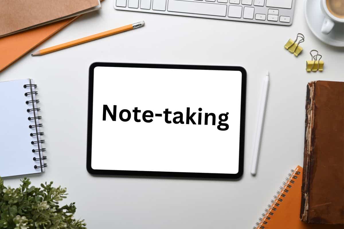 A desk with a notebook, keyboard, and iPad with stylus (with the word note-taking)