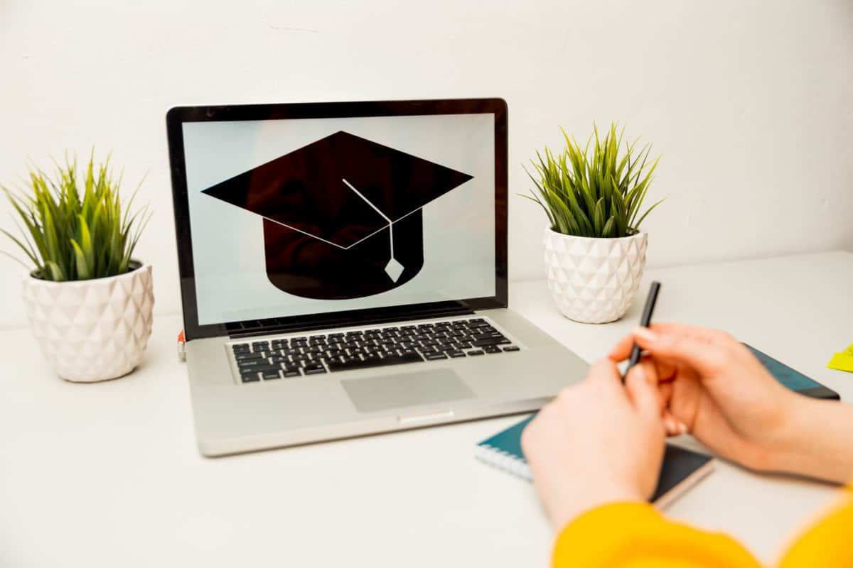 Image of a graduation cap on a laptop screen. The laptop is sitting on a desk in front of a woman's hands holding a notebook and pen.